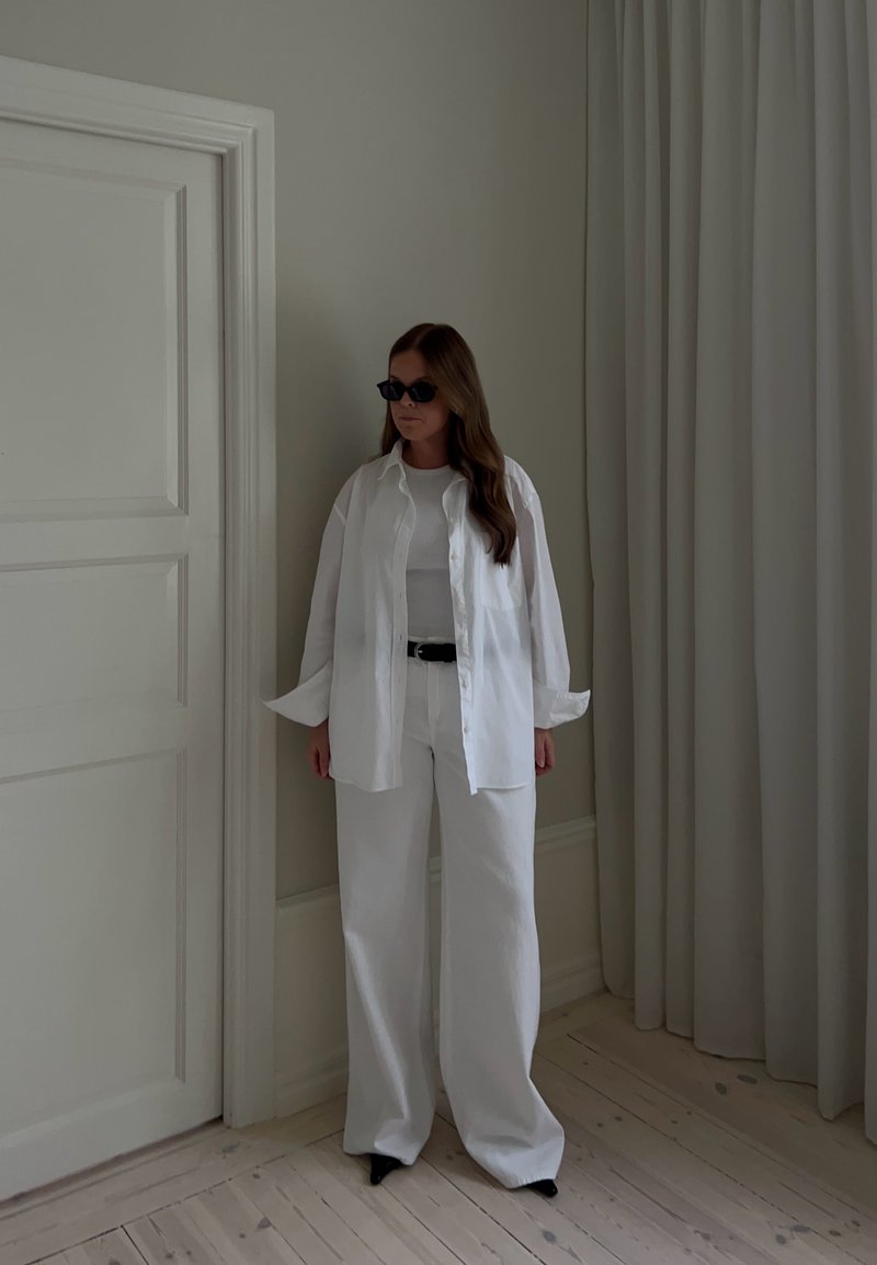 Femme portant une chemise blanche oversize, un pantalon blanc à jambes larges, une ceinture noire et des lunettes de soleil noires, debout à l'intérieur près d'une porte fermée et de rideaux.