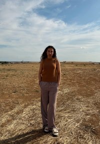 Top senza maniche marrone a maglia, pantaloni a gamba larga a righe e sandali bianchi. In piedi in un campo secco sotto un cielo blu con nuvole sparse.