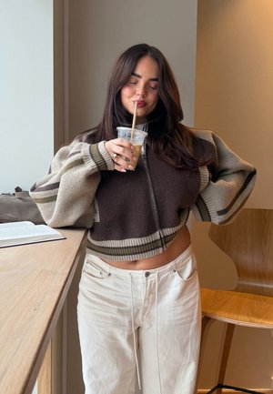 Joven mujer con cabello oscuro y largo, vistiendo un suéter corto de rayas y pantalones blancos, bebiendo café helado con los ojos cerrados junto a una mesa de madera.