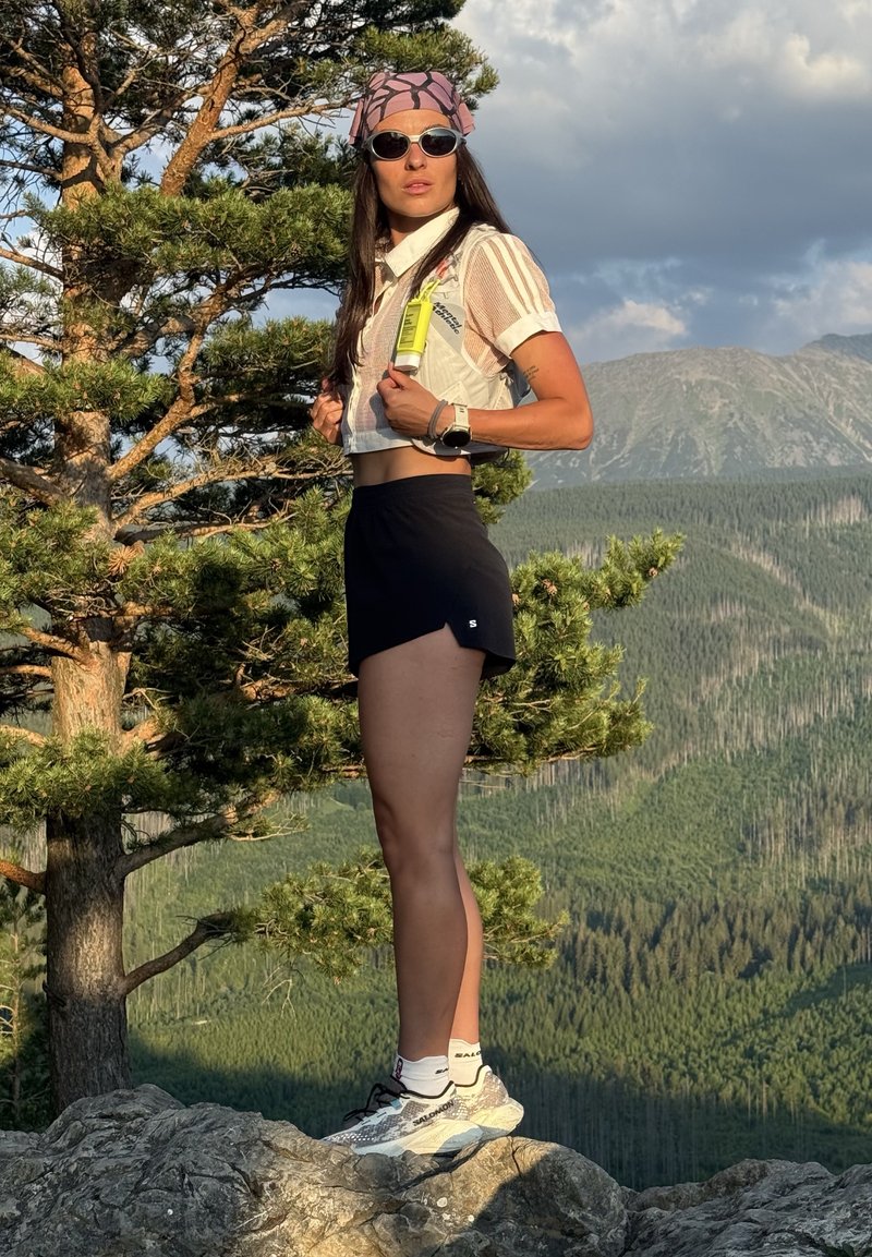Woman wearing sunglasses and a headscarf stands on a rock with mountains and pine trees in the background during daylight.