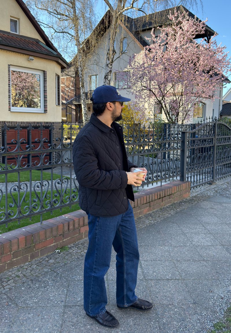 Homme portant une casquette bleu marine et une veste se tient sur un trottoir en tenant une tasse, avec des cerisiers en fleurs et des maisons derrière une clôture en fer noir.