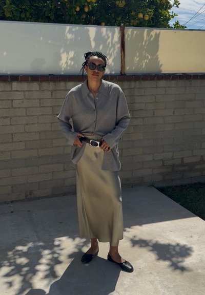 Mujer con gafas de sol, camisa gris oversize, falda beige de satén y zapatos planos negros, de pie al aire libre contra una pared de ladrillos con un limonero arriba.