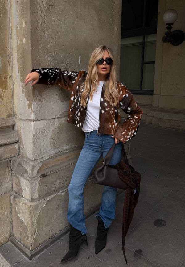 Mujer con gafas de sol, chaqueta de piel marrón con manchas, camisa blanca, jeans azules y botas negras, apoyada contra una pared de piedra, sosteniendo una bolsa negra.
