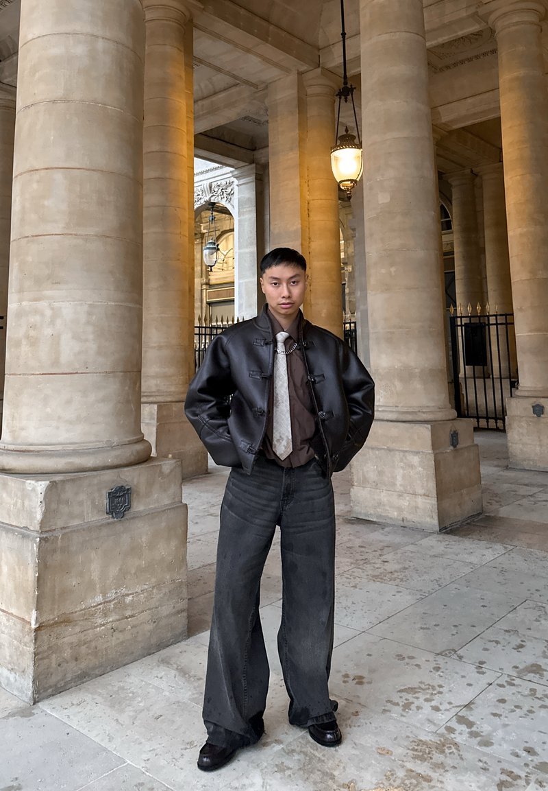 Un bărbat tânăr stă într-o colonadă arhitecturală purtând blugi largi de culoare închisă, o geacă neagră de piele, o cămașă maro, o cravată deschisă cu model și pantofi negri.