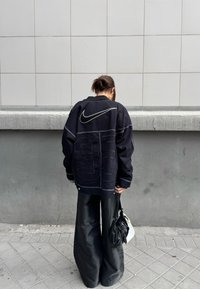 Personne debout contre un mur gris portant une veste noire surdimensionnée avec un grand logo Nike et tenant deux sacs à main.