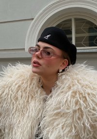 Φουντωτό λευκό ψεύτικο παλτό από γούνα, μαύρη μπερέ, ροζ γυαλιά ηλίου, και σκουλαρίκια σε σχήμα καρδιάς. Υφή, στρώσεις και σύγχρονος σχεδιασμός.