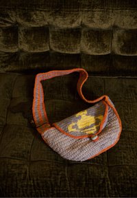 Sac à bandoulière crocheté avec bordure orange et motif de planète jaune sur le rabat, posé sur un canapé texturé brun foncé.