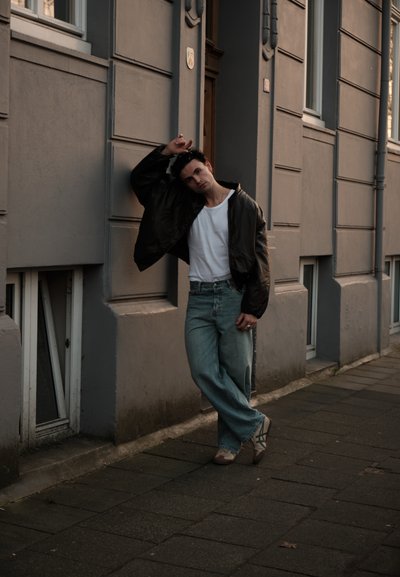 Joven con chaqueta negra, camisa blanca y vaqueros apoyado contra la pared de un edificio gris en una acera de la ciudad al anochecer.