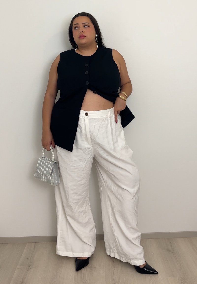 Black sleeveless top with front buttons, white linen wide-leg pants with button detail, black pointed-toe heels, and a patterned gray handbag.