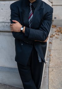 Completo grigio scuro con giacca strutturata, indossato sopra una camicia nera. Presenta una cravatta a righe rossa e grigia. Orologio argento al polso.