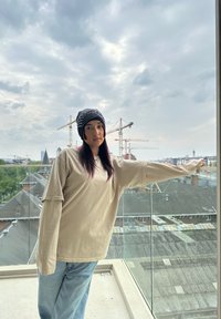 Beige longslavige shirt met brede mouwen, denim jeans en een gestreepte zwarte beanie. Op de achtergrond zijn kranen en daken te zien onder een bewolkte lucht.