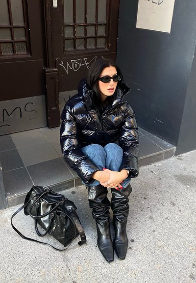 Chaqueta acolchada negra con textura brillante, vaqueros de mezclilla azules, botas negras holgadas y un bolso negro con asas sobre una superficie de hormigón.