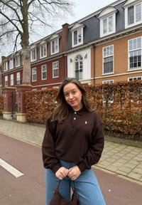 Jeune femme debout dans une rue devant des haies brunes et des bâtiments en briques, portant un pull brun foncé et un jean bleu clair tout en tenant un sac.