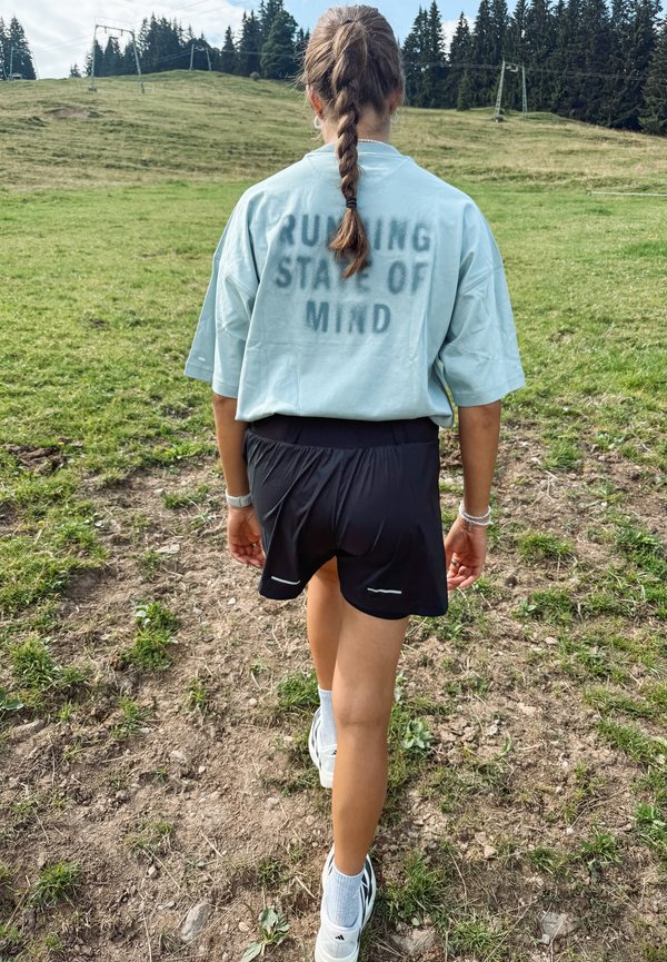 Junge Frau mit Zopf geht auf einem grasbewachsenen Weg, trägt Shorts und ein Shirt mit der Aufschrift "RUNNING STATE OF MIND", mit Wald im Hintergrund.