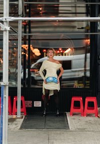 Triibuline kollane kleit asümmeetrilise äärega, mustad sukkpüksid ja pahkluu saapad. Hoides sinist pilvemustrilise kotiga, seisab restorani ees.