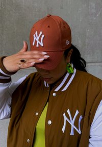 Ruskea baseball-lippis, jossa on valkoinen NY-logo, käytetty ruskean ja valkoisen yliopistotakin kanssa. Näkyvissä vihreä yläosa ja suuret vihreät korvakorut.