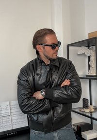 Blouson aviateur en cuir noir avec un col montant, des boutons-pression et des poignets côtelés. Porté avec des lunettes de soleil foncées et un jean en denim.