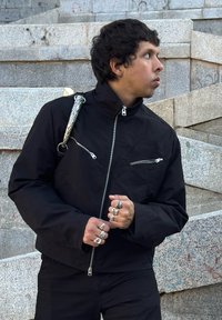 Black jacket with silver zippered pockets, paired with black pants. Features textured fabric, a stand collar, and a decorative strap detail.