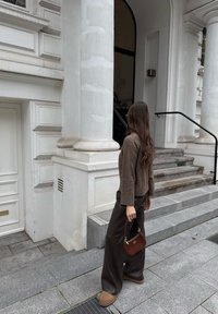 Rjav pleteni pulover, temne široke hlače, rjava torbica in svetlo rjavi škornji. Stane blizu bele zgradbe s stebri in kamenimi stopnicami.