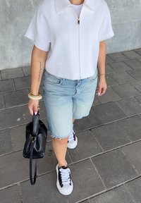 Top cropped bianco con zip, bermuda in denim azzurro chiaro, borsa nera e sneaker in bianco e nero con dettagli texture.