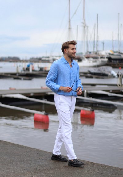 Camisa de lino azul claro con botones, pantalones blancos y zapatos negros. El fondo incluye barcos y un entorno de costa.