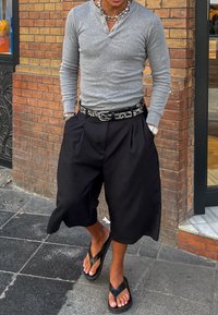 Grå langermet skjorte, svarte plisserte shorts, svart belte med sirkulære aksenter, og svarte flipe-flops. Murvegg bakgrunn.