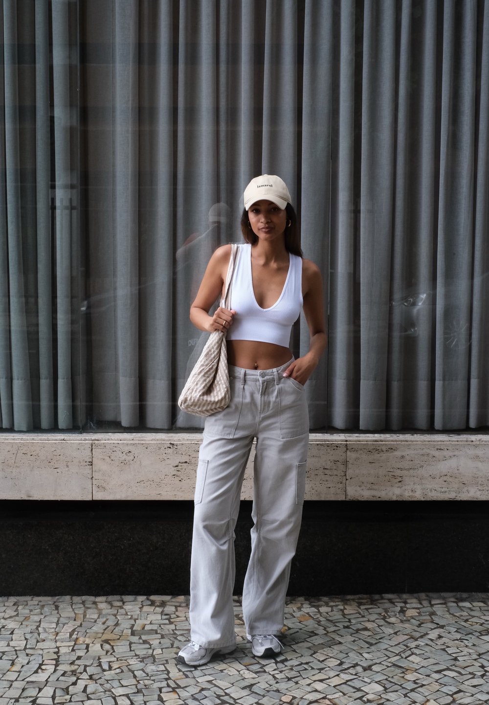 Fehér bordázott cropped top, világosszürke cargo nadrág, bézs sapka és mintás vászontáska. Mosaik csempepadlón áll, háttérben szürke függönyök.