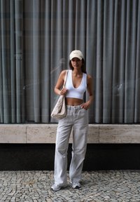 Fehér bordázott cropped top, világosszürke cargo nadrág, bézs sapka és mintás vászontáska. Mosaik csempepadlón áll, háttérben szürke függönyök.