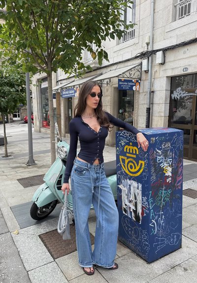 Blusa de botones de manga larga color marino, jeans de pierna ancha azul claro, sandalias negras, sosteniendo una bolsa gris, con una caja de utilidad azul cubierta de graffiti cerca.