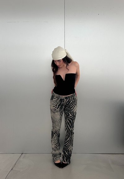 Mujer con top negro sin tirantes, pantalones con estampado de cebra, tacones negros puntiagudos y un pañuelo crema en la cabeza, de pie frente a una pared gris lisa.