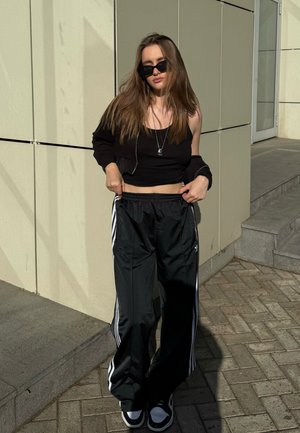 La ropa deportiva negra incluye un top corto ajustado y pantalones sueltos con bandas laterales blancas. Unas gafas de sol negras y zapatillas completan el look.