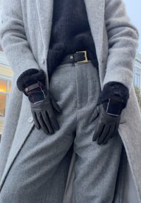 Personne portant un manteau en laine gris et un pantalon, un pull noir, une ceinture noire et des gants en cuir noir avec des poignets à carreaux en extérieur.