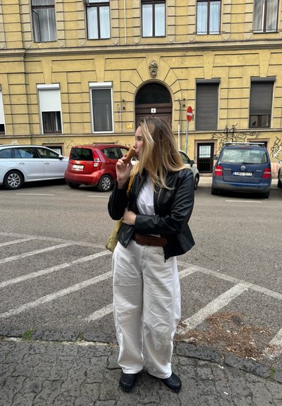 Una persona que lleva una chaqueta de cuero negra y pantalones anchos blancos sostiene un cucurucho de helado, de pie en una calle bordeada de coches estacionados.