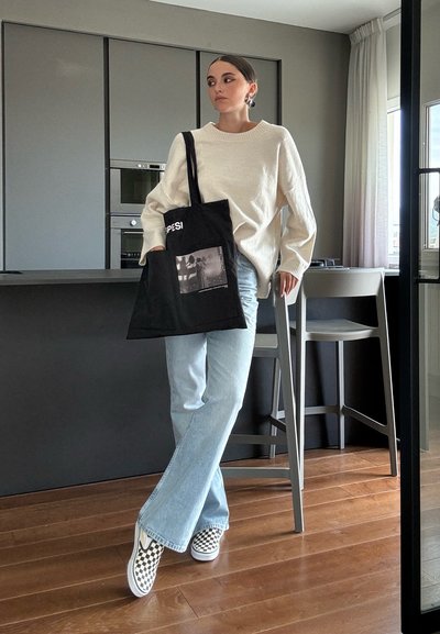 Bolsa tote negra con un bolsillo para fotos y texto en blanco, suéter crema, jeans acampanados de color azul claro y zapatillas de cuadros en blanco y negro.
