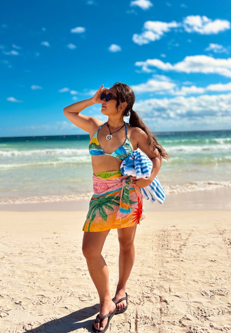 Mujer con gafas de sol, bikini colorido y pareo, sosteniendo una toalla a rayas, de pie en una playa de arena con el océano y el cielo azul de fondo.