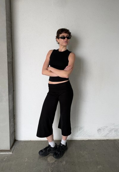 Persona con cabello corto y rizado, que lleva una camiseta negra sin mangas, pantalones anchos negros, zapatillas negras, calcetines blancos y gafas de sol oscuras, de pie con los brazos cruzados.
