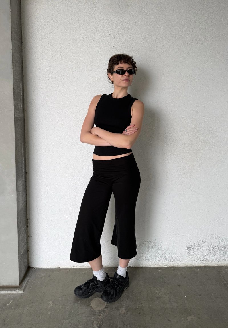 Personne aux cheveux courts et bouclés portant un haut sans manches noir, un pantalon large noir, des baskets noires, des chaussettes blanches et des lunettes de soleil foncées, debout les bras croisés.