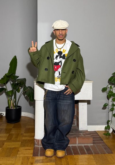 Abrigo verde de doble botonadura, camiseta blanca con gráfico de Mickey Mouse, vaqueros azules holgados, calzado marrón claro y detalles en cadena dorada.