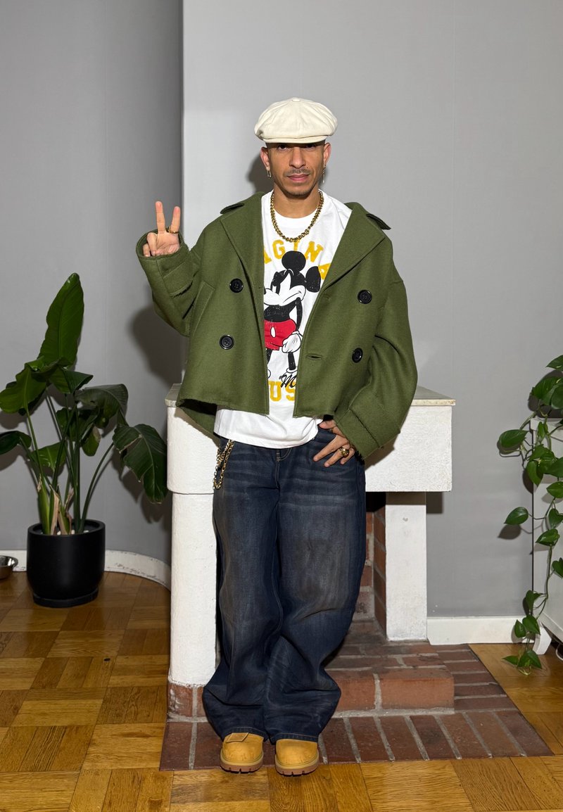 Cappotto verde doppiopetto, maglietta bianca con grafica di Topolino, jeans blu larghi, calzature color cuoio e dettagli con catena d'oro.