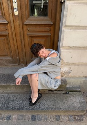 Persona con cabello rizado corto que lleva una sudadera con capucha gris y pantalones cortos, sentada en unos escalones de piedra frente a una puerta de madera.