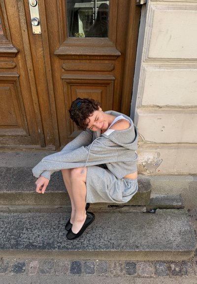 Persona con cabello rizado corto que lleva una sudadera con capucha gris y pantalones cortos, sentada en unos escalones de piedra frente a una puerta de madera.