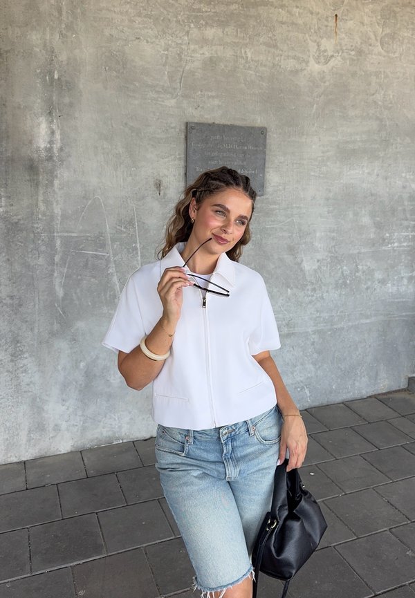 Top bianco a maniche corte con zip, pantaloni corti in denim chiaro con orlo strappato, tiene occhiali da sole neri, accessoriate con un bracciale spesso e una borsa piccola.