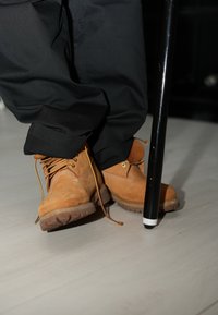 Braune Lederstiefel mit gelben Schnürsenkeln, robusten Sohlen und seitlicher Naht; schwarzer Gehstock mit weißer Spitze; auf hellem Holzboden ruhend.