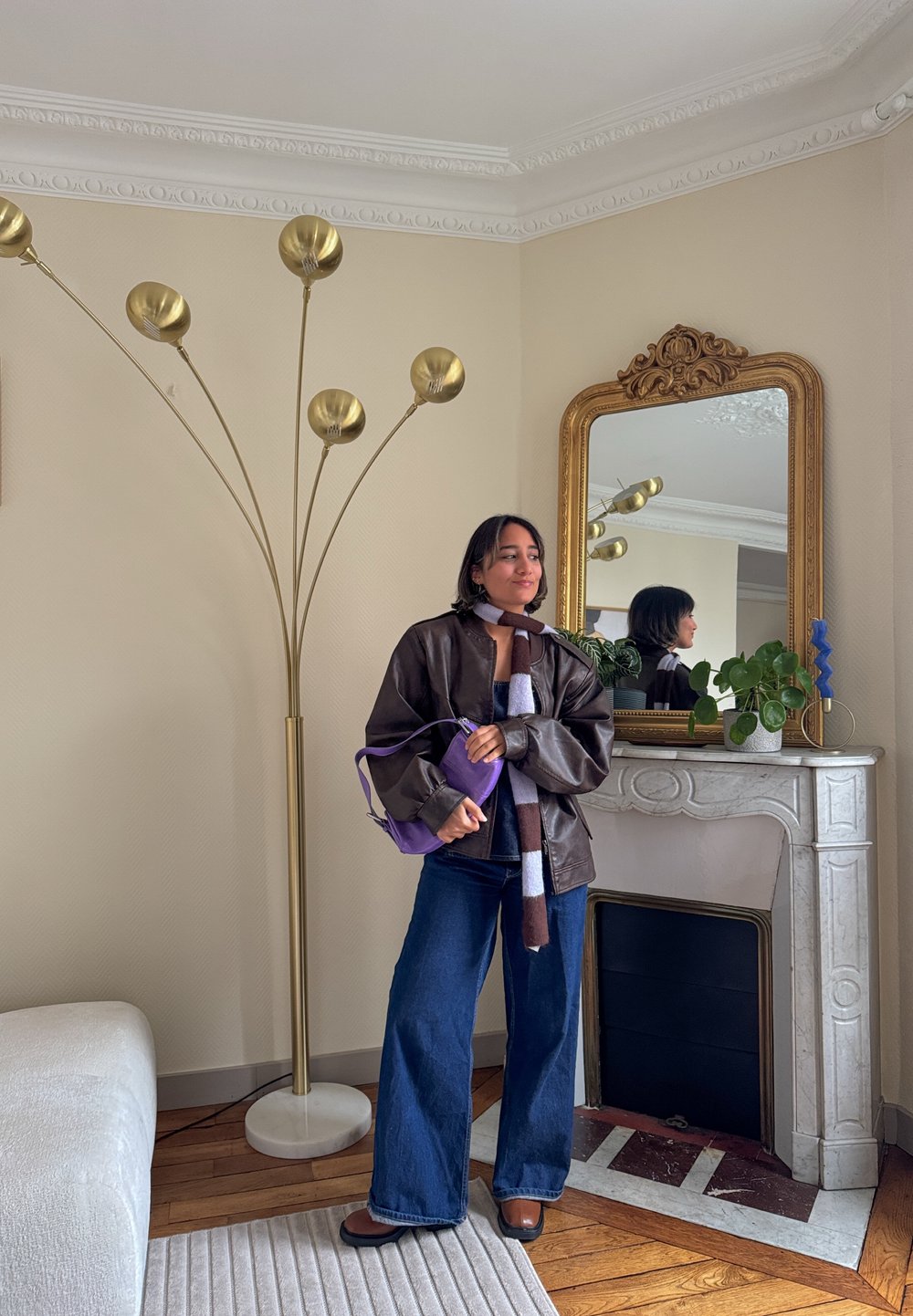 Veste en cuir marron, écharpe rayée, pantalon en denim évasé, sac à main violet. La pièce comprend un lampadaire doré, un miroir et une plante en pot.