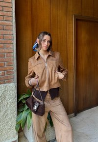 Brązowa zamszowa kurtka z ozdobnymi ćwiekami, zestawiona z ciemnobrązową bluzką i spodniami. Brązowa torebka ze sztruksu o prostym kroju. W tle zielone rośliny.