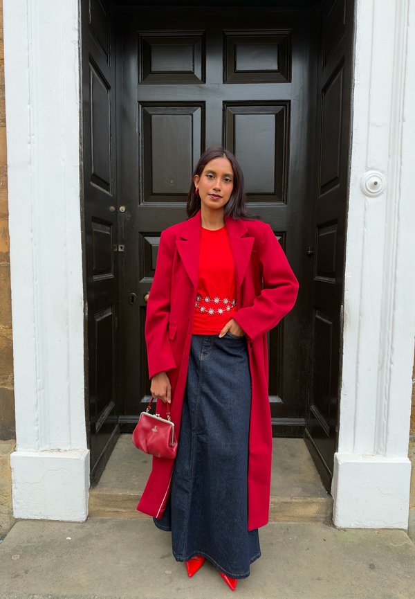 Une femme pose devant une entrée noire. Elle porte un manteau et un haut rouges, une longue jupe en jean et tient un sac à main rouge.