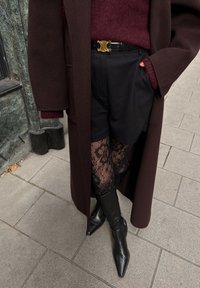 Dunkelbrauner Mantel über einem kastanienbraunen Pullover, schwarze Shorts und Spitzenstrumpfhosen. Schwarze Stiefel mit spitzer Spitze und einem goldenen Gürtel mit rundem Design.