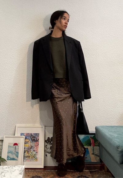 Joven con cabello largo y oscuro recogido lleva una blazer negra de gran tamaño, suéter oliva y falda marrón brillante con puntos blancos, de pie en el interior.