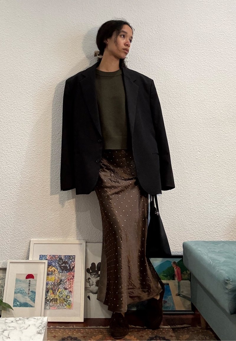 Jeune personne aux longs cheveux noirs attachés porte un grand blazer noir, un pull olive et une jupe marron brillante à pois blancs, debout à l'intérieur.