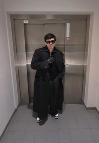 Fekete hosszú kabát, fekete pulóver, vékony csíkos nadrág, fehér cipő fekete talppal, és fekete kesztyűk. Napszemüveg, a lift közelében áll.